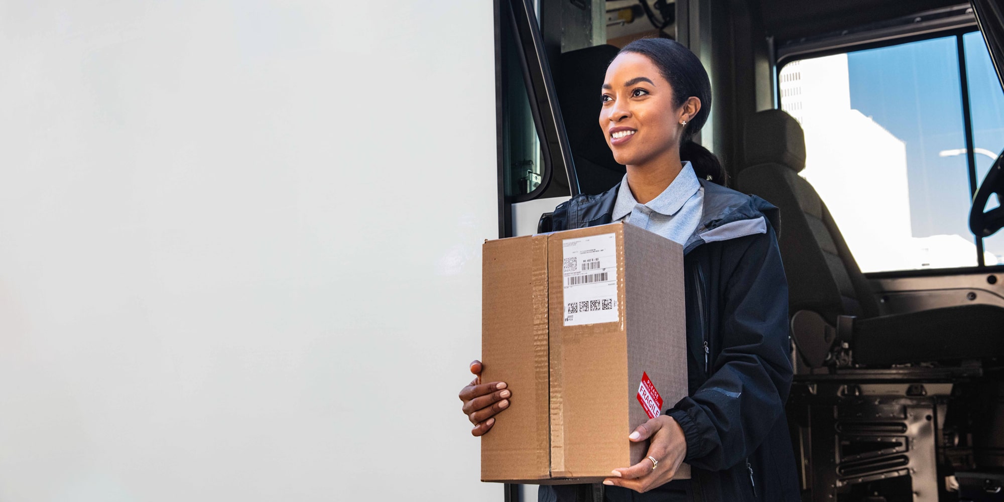 A delivery driver carrying a package and exiting a BrightDrop Zevo delivery vehicle.