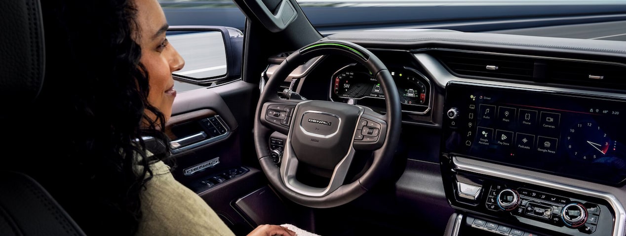 A profile view of a woman with curly dark hair utilizing the hands-free driving feature, Super Cruise, on a GMC vehicle, the interior is modern with a multi-function steering wheel that has the DENALI logo at its center and a large digital dashboard and infotainment screen.