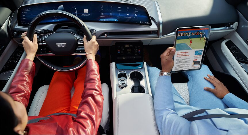 An overhead view of two people in the front seats of a Cadillac, with the driver in a red jacket holding the steering wheel and the passenger in a blue shirt and pants holding a tablet displaying a live sports game.