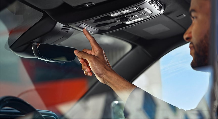 A man with a beard is reaching up to press the OnStar button on the overhead console of a vehicle, the rearview mirror and front windshield are also visible.