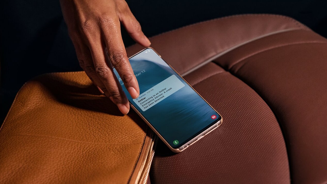 A person's hand reaches for a rose-gold smartphone resting on a brown leather handbag, displaying an OnStar notification that reads "OnStar Emergency Advisor has detected a crash."