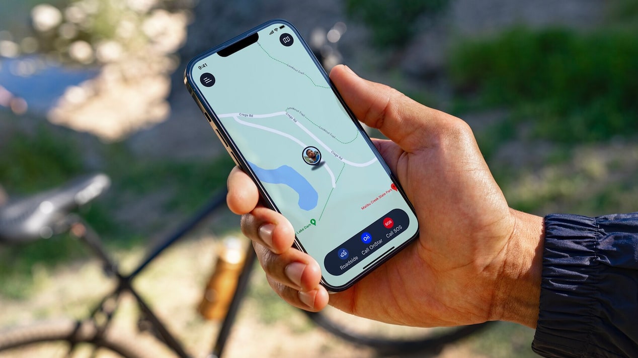 A hand holds a smartphone displaying a map application with the user's location near a lake, showing options to "Roadside," "Call OnStar," and "Call SOS," with a bicycle blurred in the background, suggesting outdoor activity.