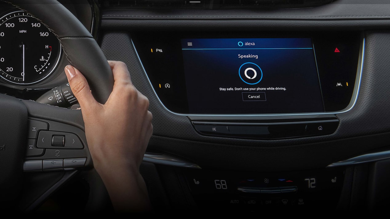 A person's hand is holding the steering wheel of a vehicle, with the infotainment screen displaying the Amazon Alexa interface in a car, showing a "Speaking" animation and a safety message: "Stay safe. Don't use your phone while driving."