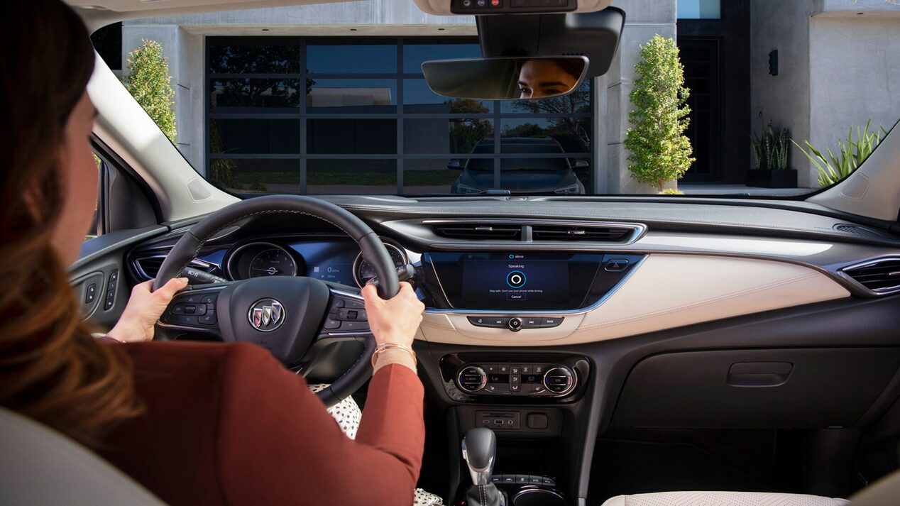 The interior view from the driver's seat of a Buick, showing a woman's hands on the steering wheel while the infotainment screen displays the Amazon Alexa "Speaking" interface, with a garage door visible through the windshield.